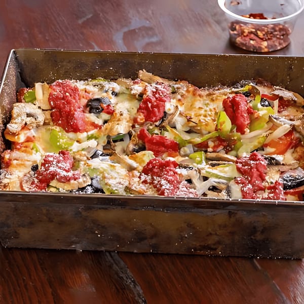 A Detroit-style pizza in a rectangular pan, topped with vegetables, cheese, and dollops of red sauce.