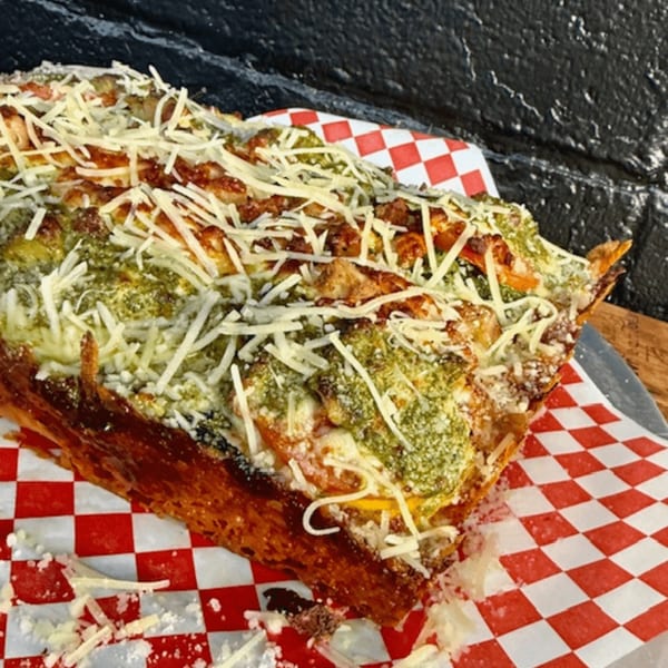 A thick-crust Detroit Pizza topped with pesto, cheese, and chicken, on red and white checkered paper.